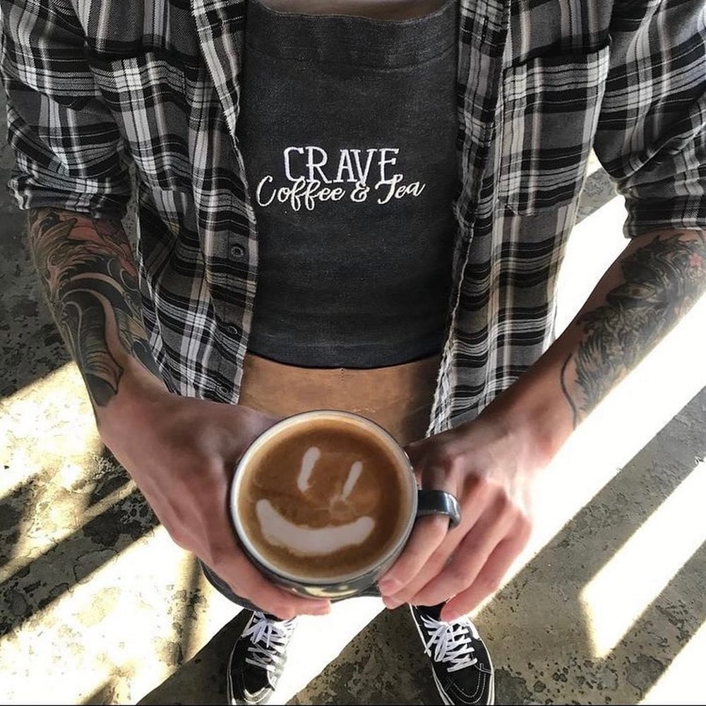 Barista in a Crave Coffee & Tea apron holding a latte with smiley latte art